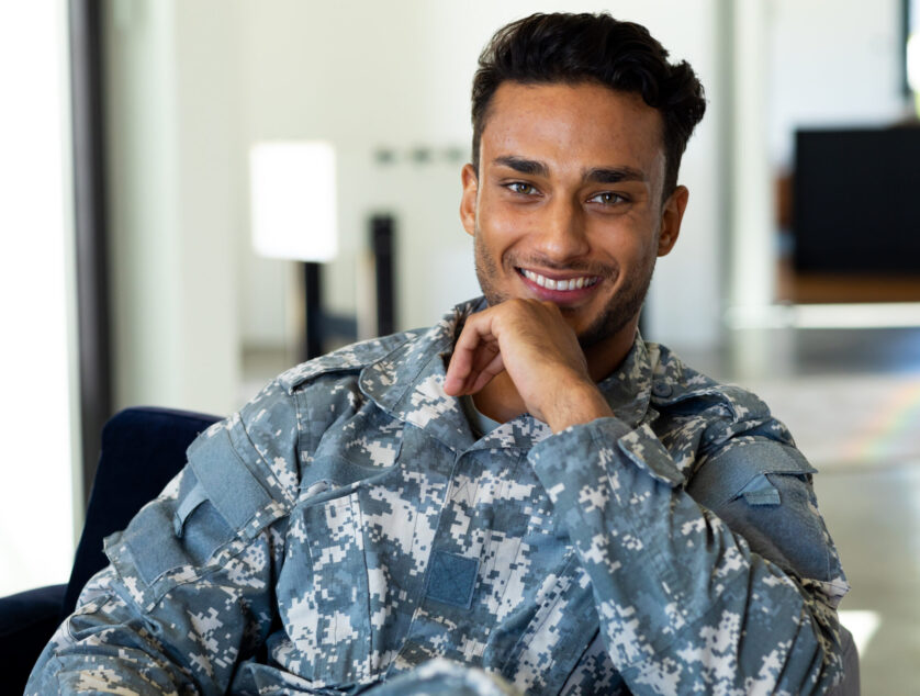 Portrait of happy biracial male soldier wearing military uniform, sitting in armchair at home. Lifestyle, military service and free time, unaltered.